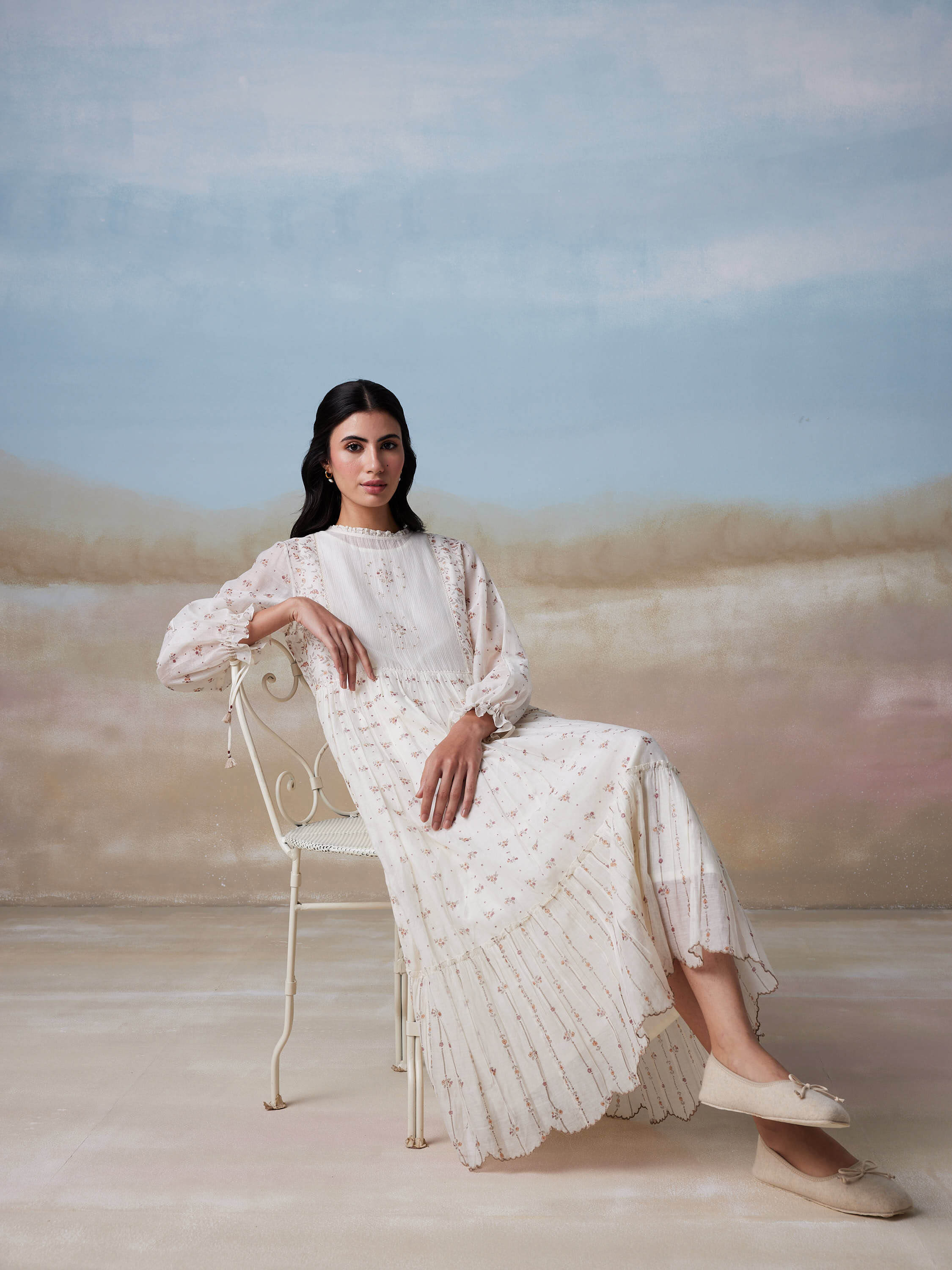 Woman in elegant white dress seated on vintage chair against pastel background.