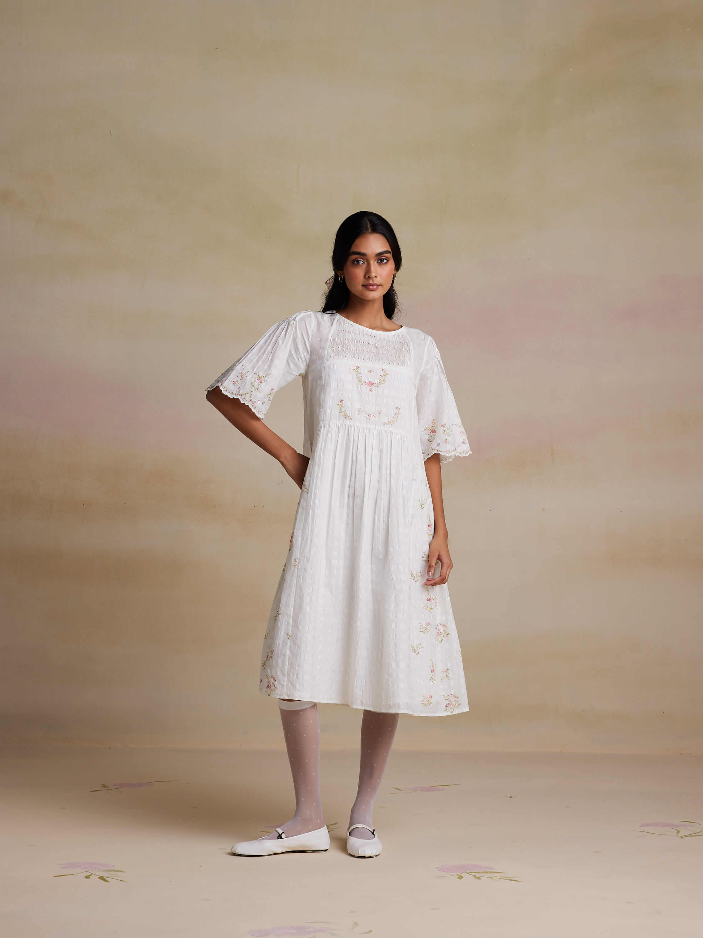 Woman in white dress with floral embroidery, posing against neutral background.