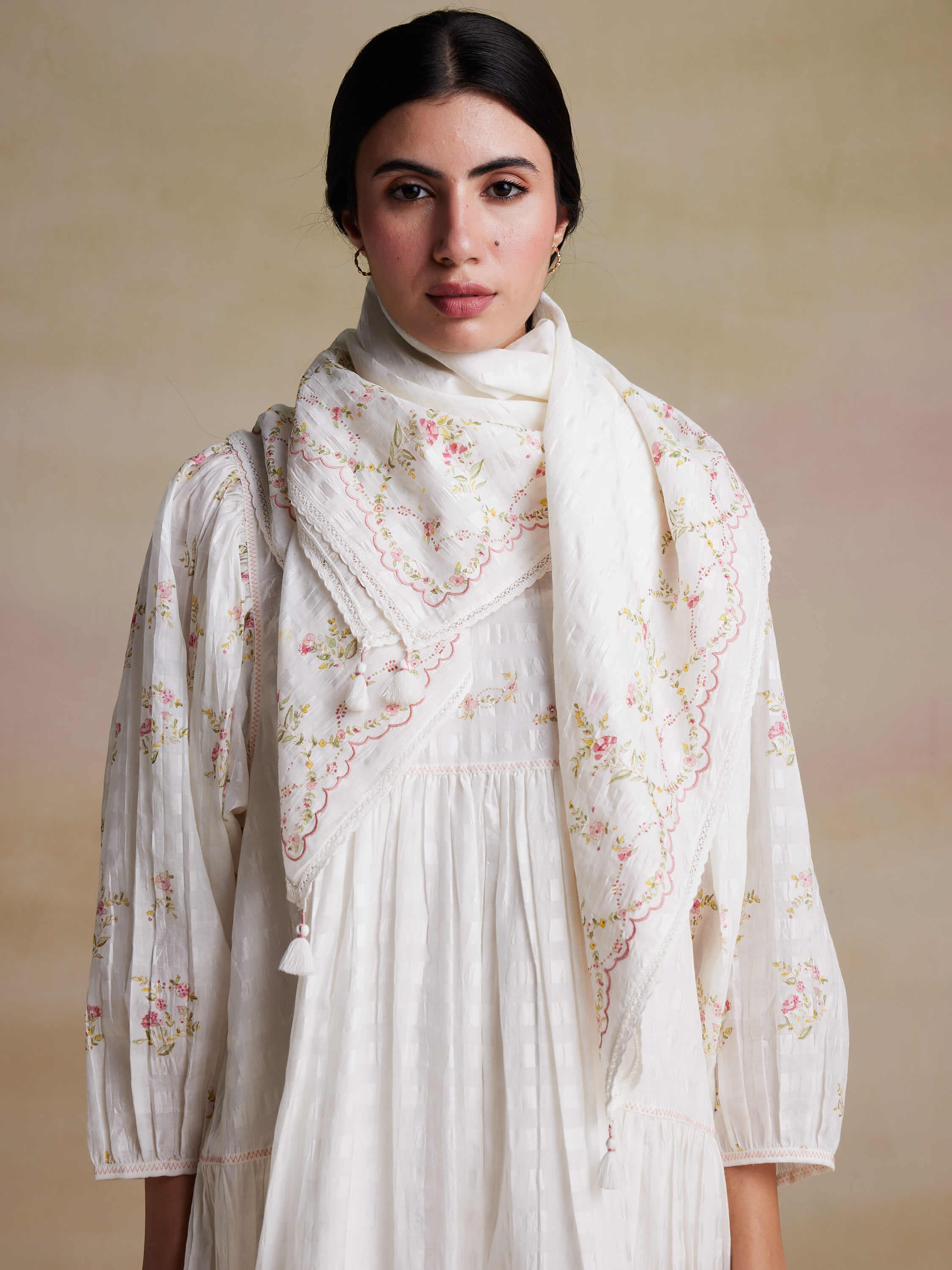 Woman in white flower-embroidered outfit and scarf against plain background.