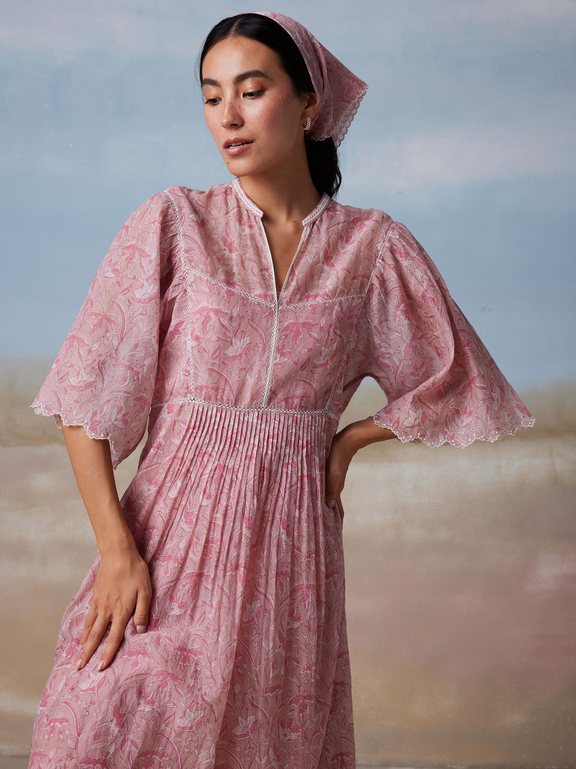 Woman in elegant pink floral dress and matching headscarf against pastel background.