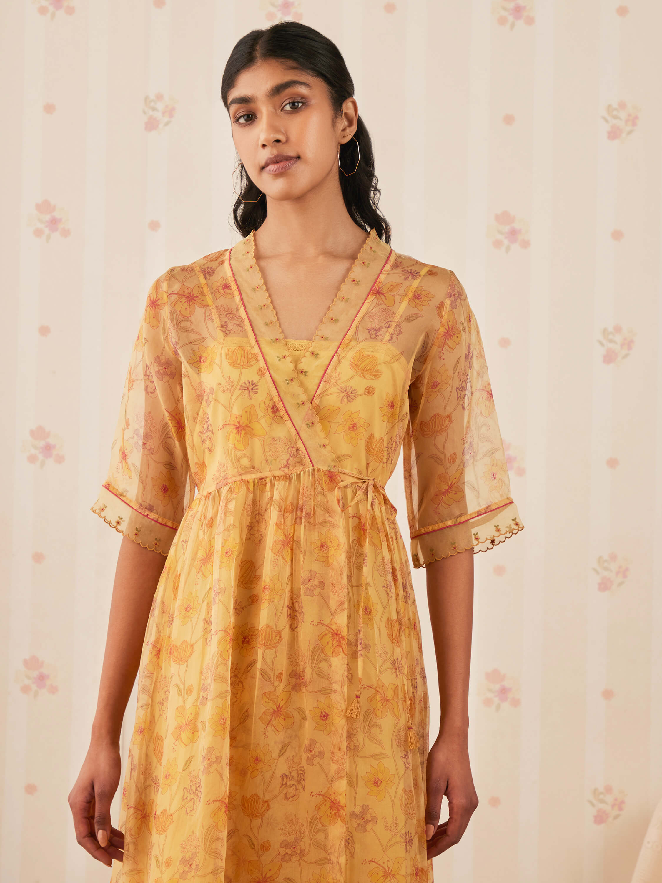 Woman in floral yellow dress with sheer sleeves standing indoors.