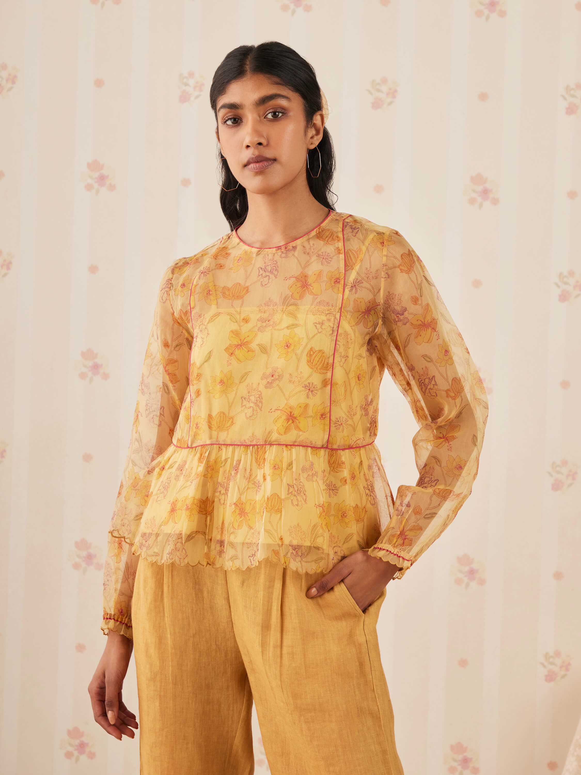 Woman in yellow floral blouse and yellow trousers against floral wallpaper.