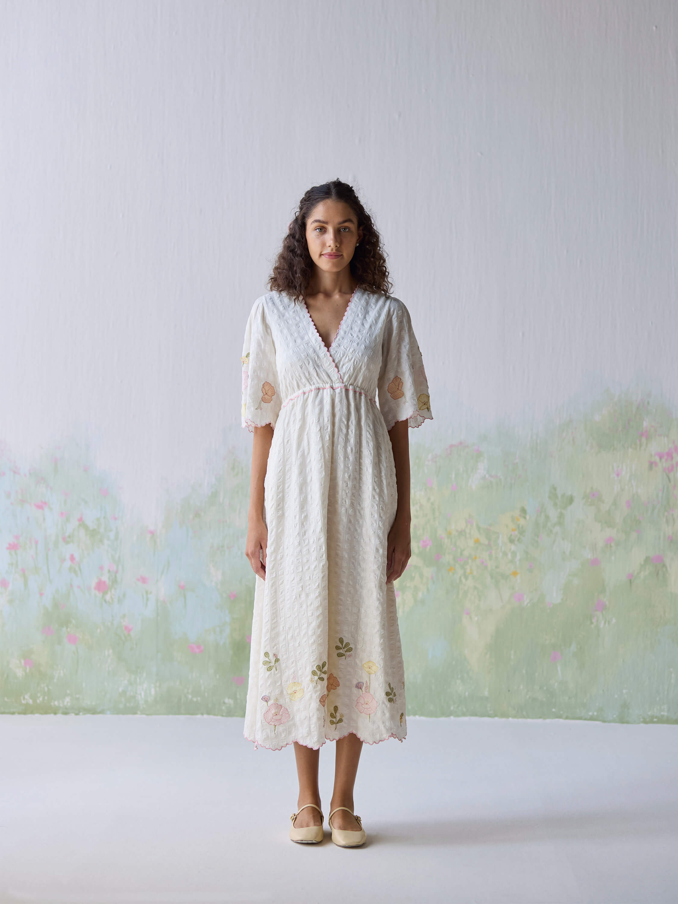 Woman wearing floral embroidered white dress standing against a pastel background.