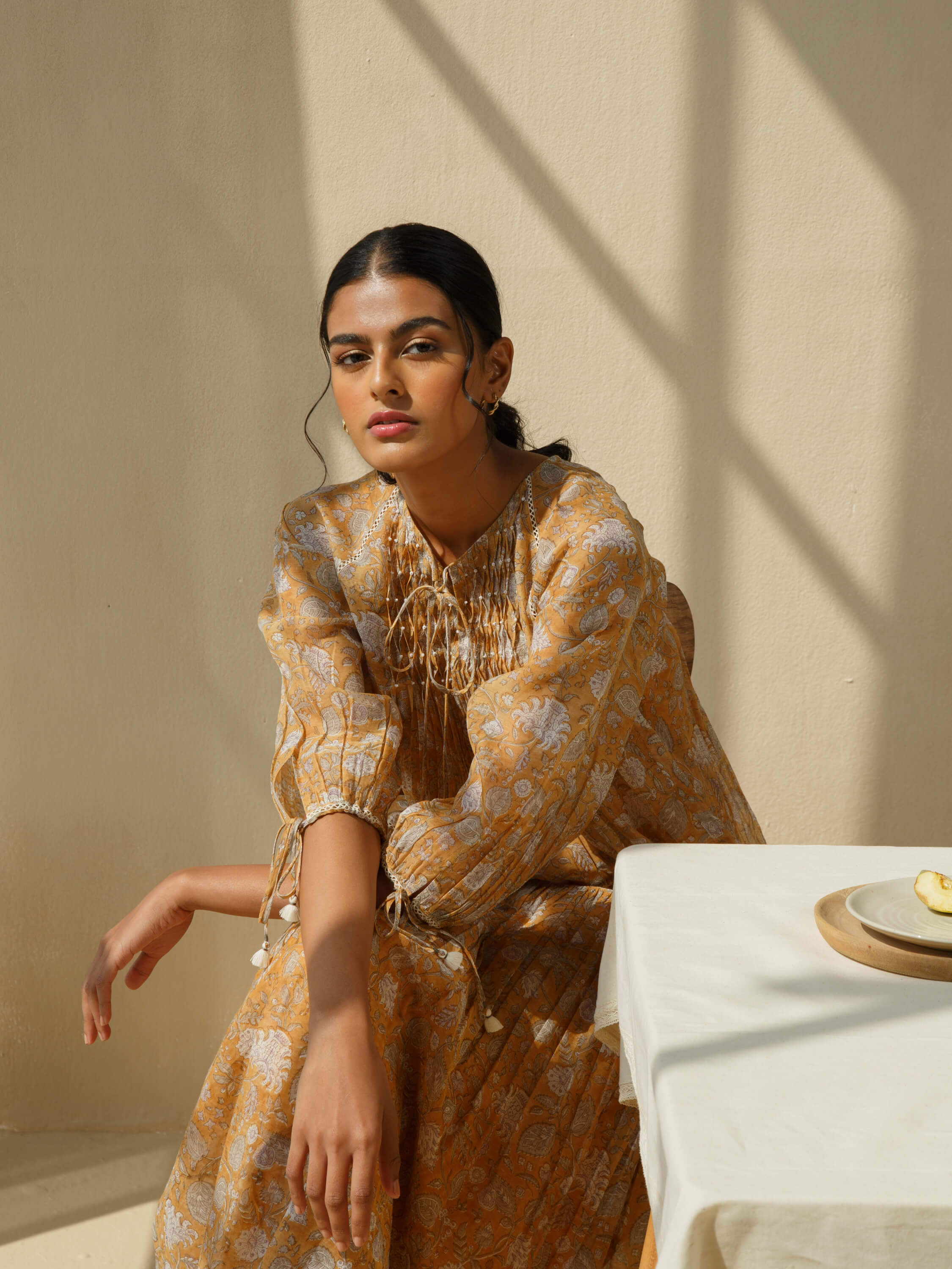 Elegant woman in mustard floral dress sitting by a table in sunlight