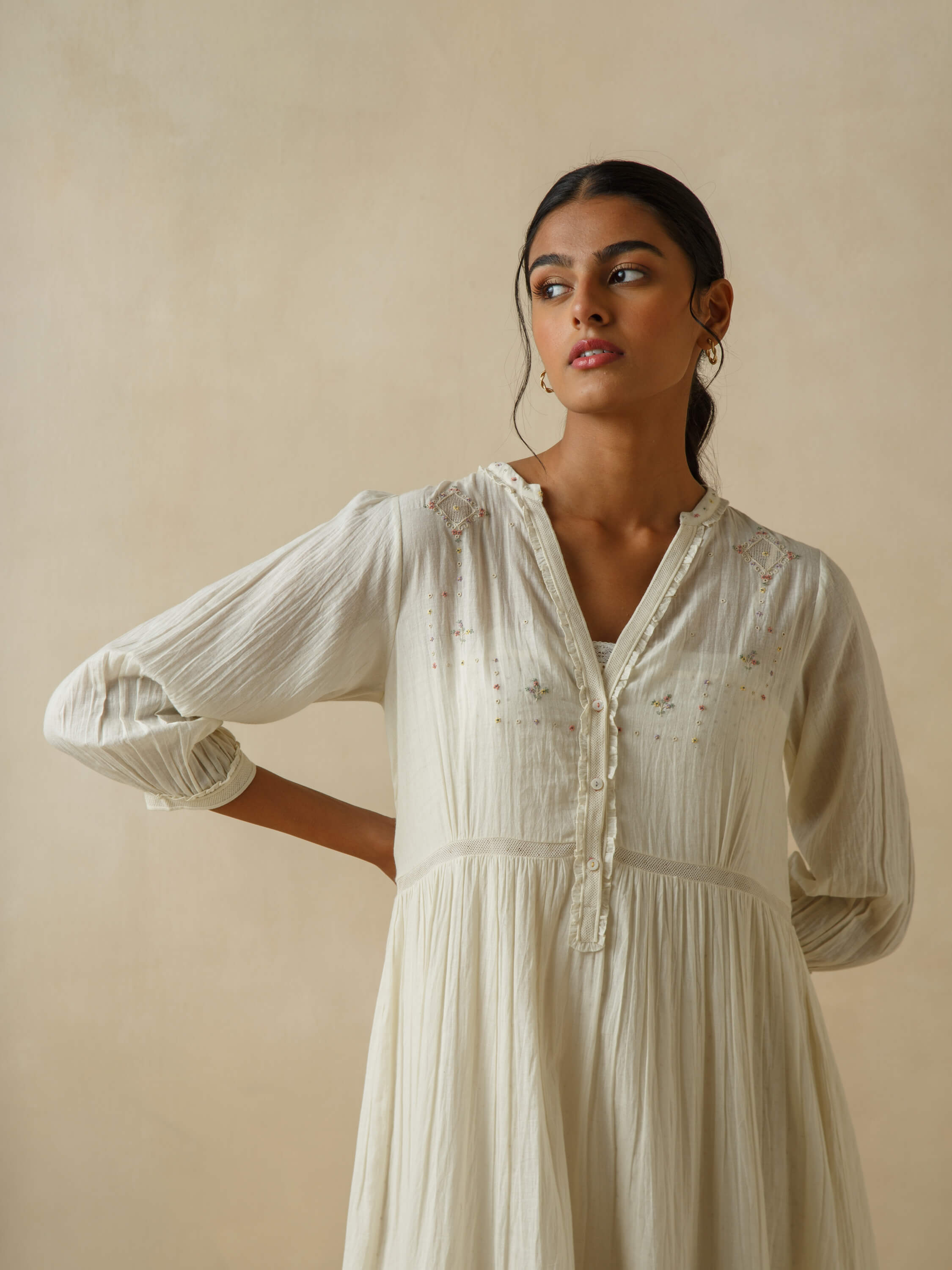 Woman in off-white embroidered dress standing against beige background.