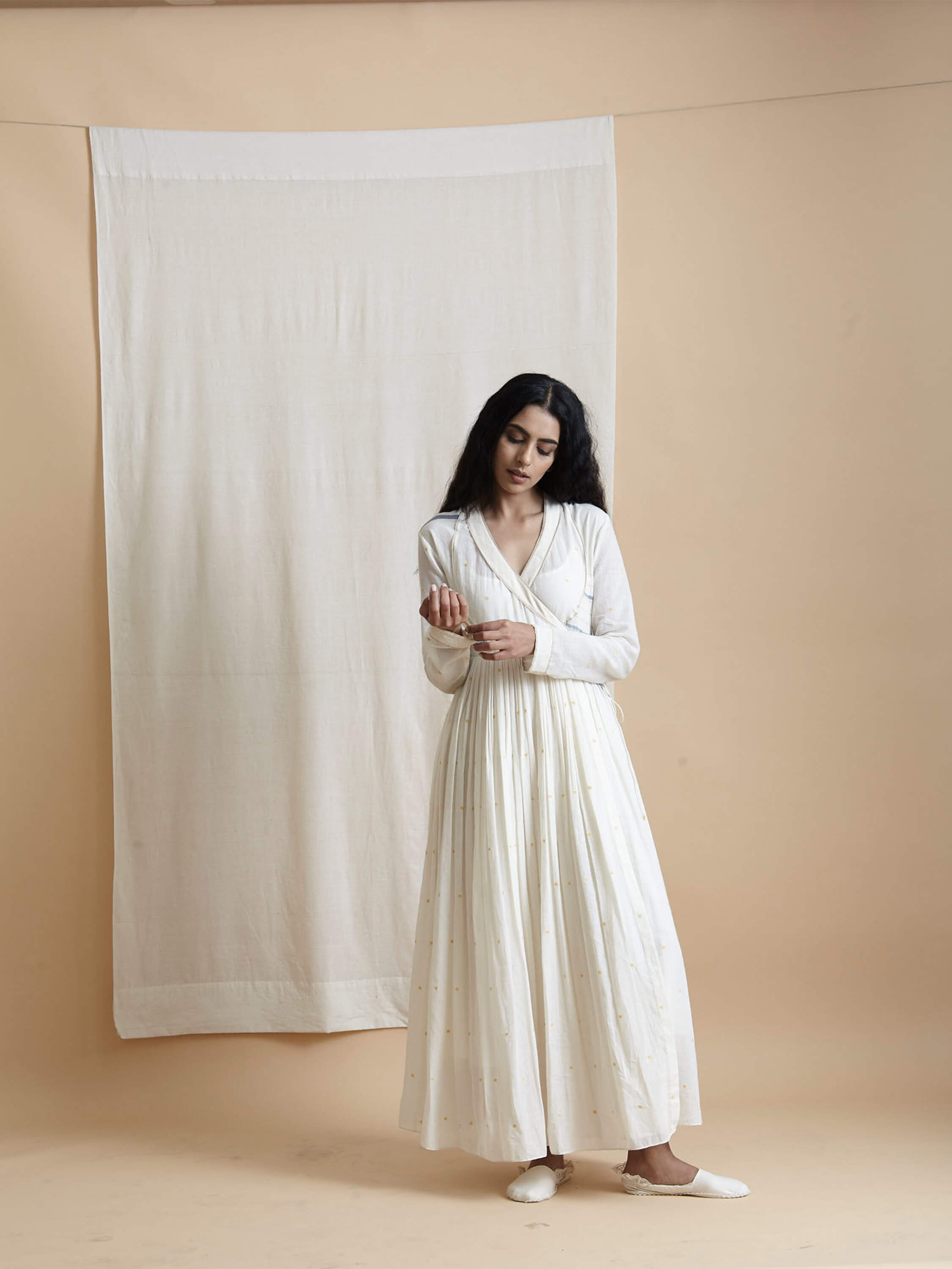 Woman in white maxi dress standing against beige background curtain.