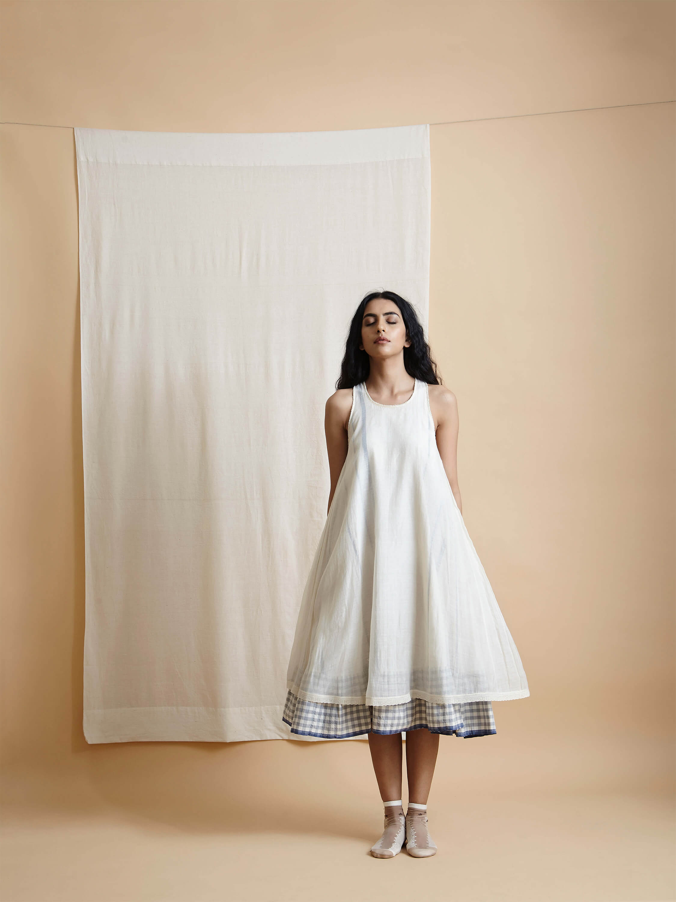 Woman in white dress standing against beige background with curtain backdrop.