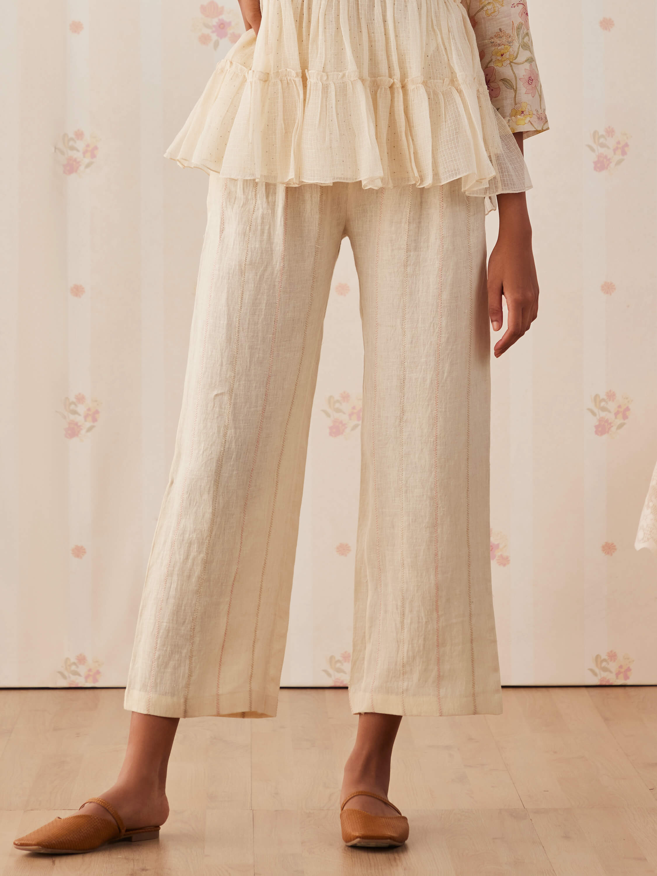 Woman in cream ruffled top and linen pants with brown flats.
