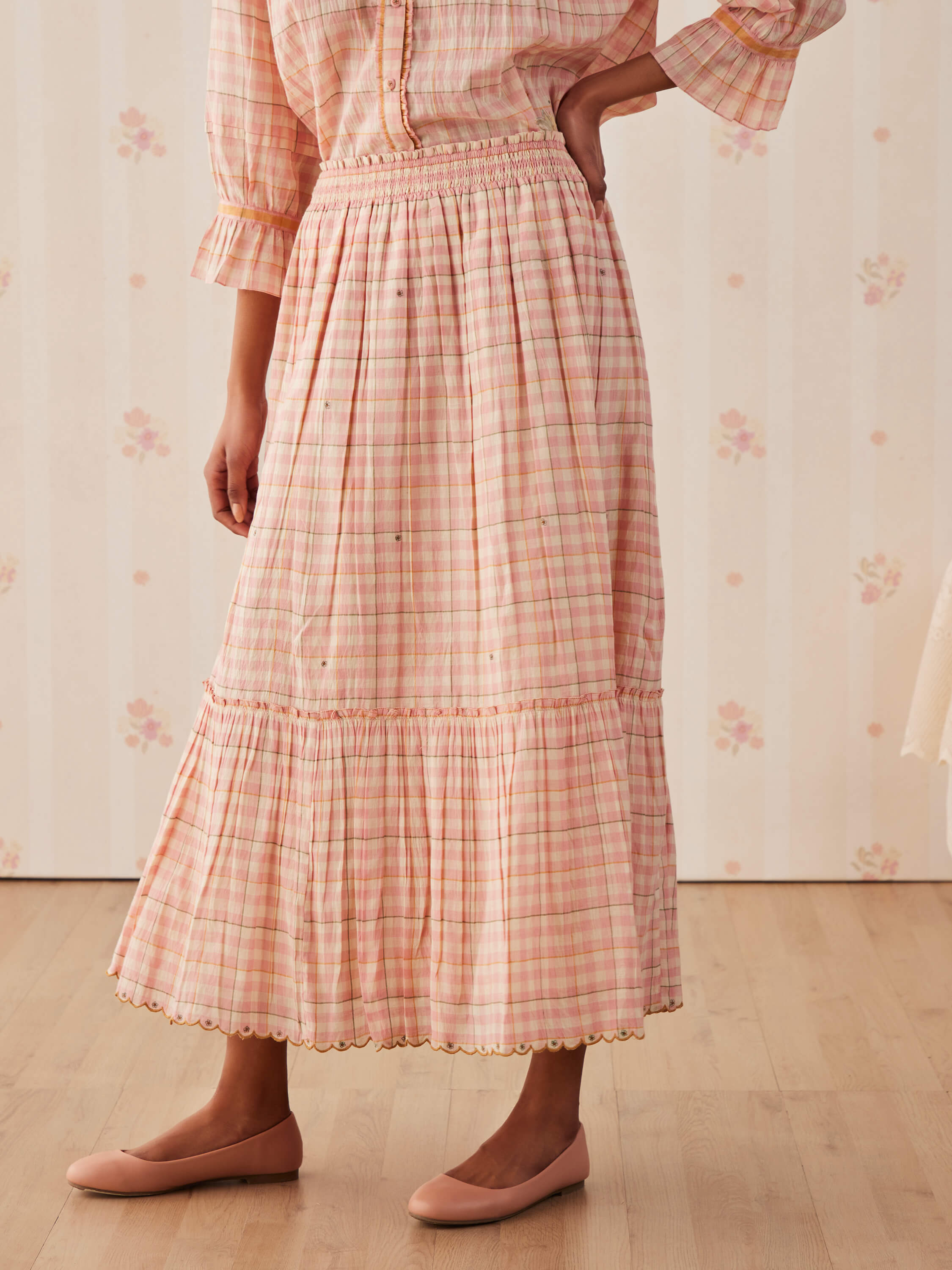 Woman wearing pink plaid midi skirt and pink flats, standing on wooden floor.