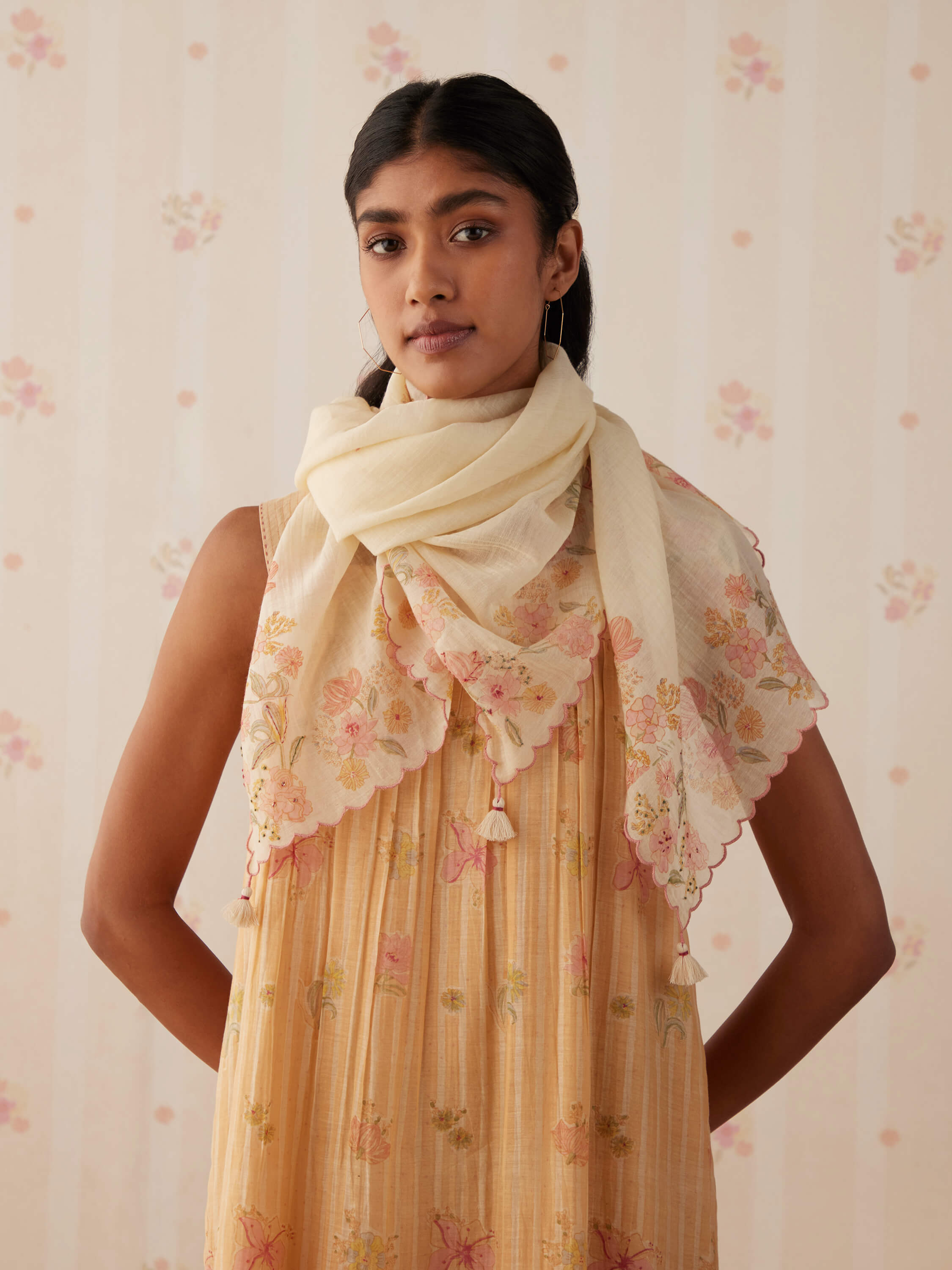 Woman in floral dress and scarf, standing against light flower-patterned wallpaper.