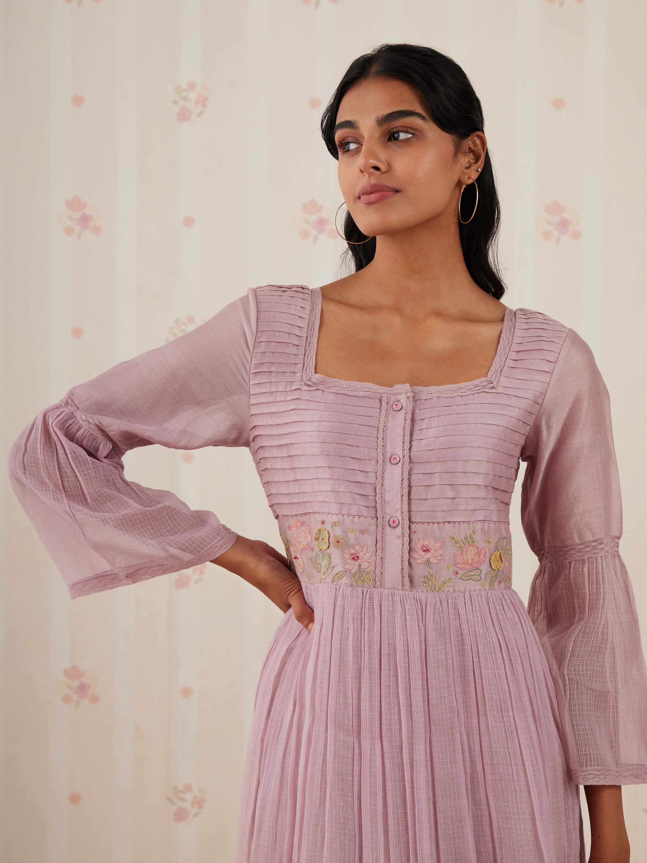 Woman in lilac pleated dress with floral embroidery and hoop earrings.