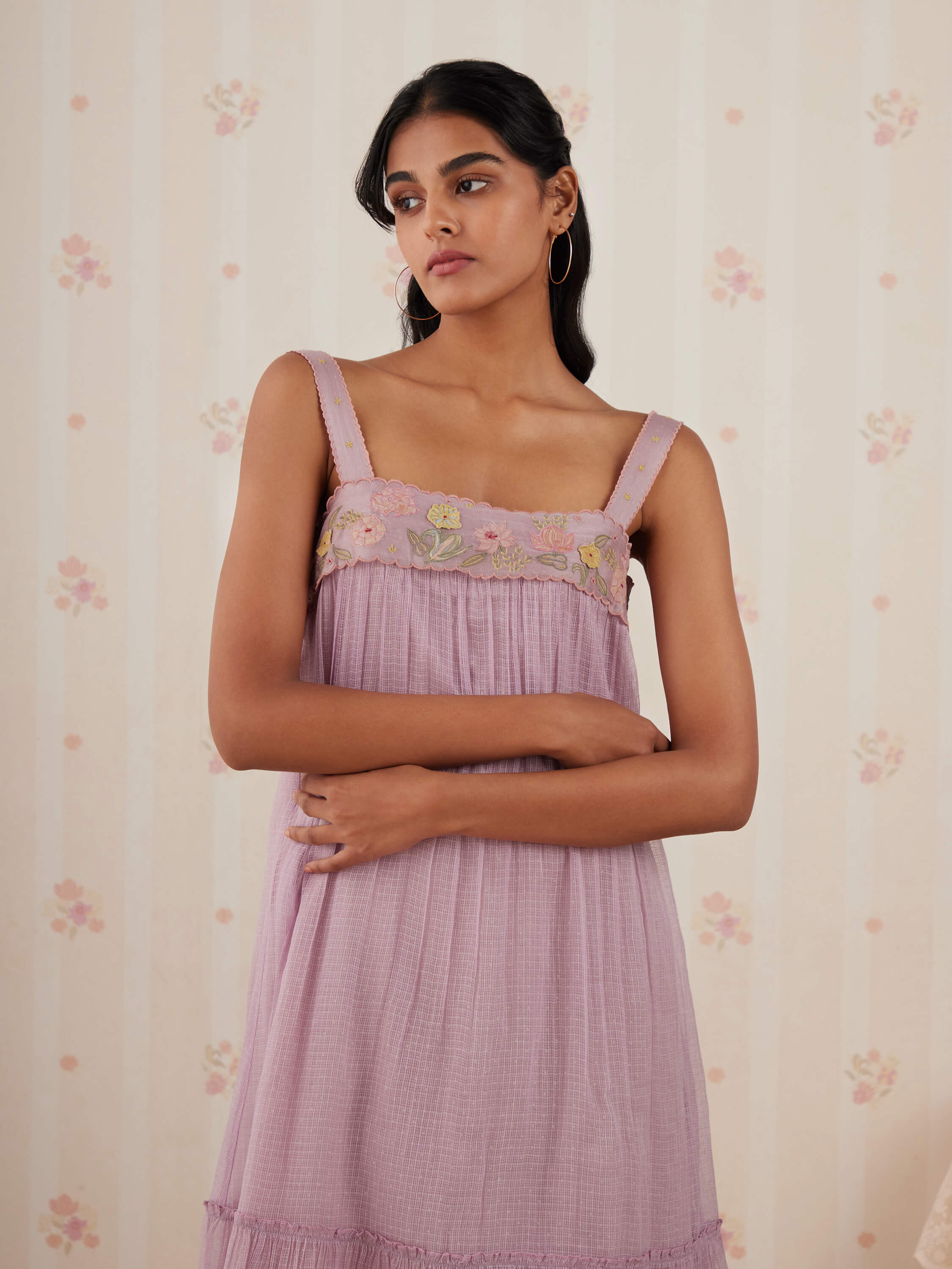 Woman in lavender dress with floral embroidery, standing against floral wallpaper.